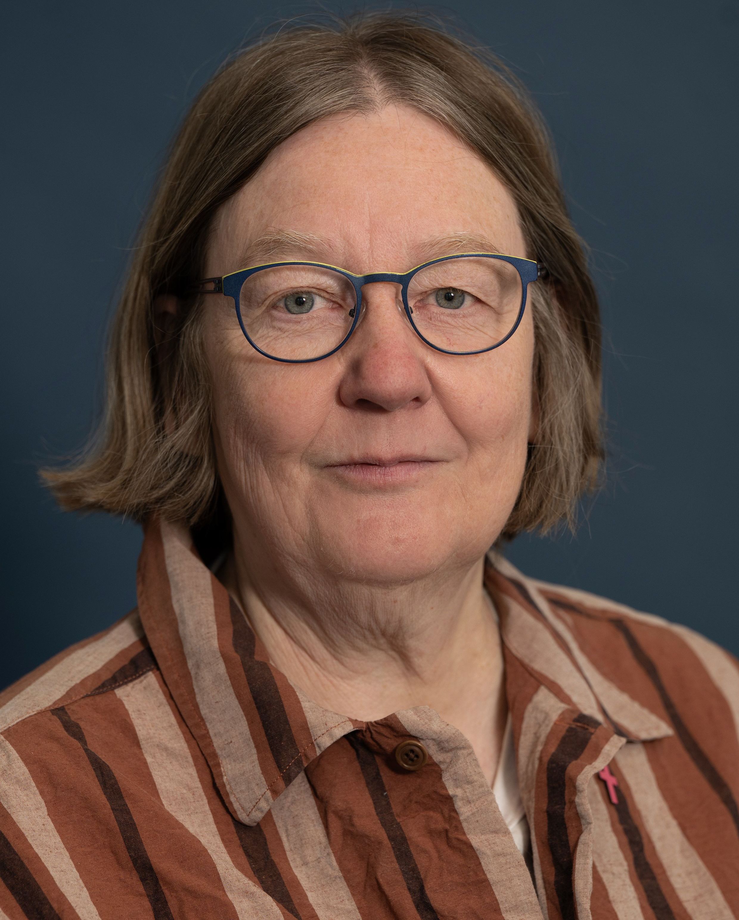 Lächelnde Frau mit runder Brille, über den Ohren geschnittenem Haar, in wollweißem Hemd, mit knubbeliger Nase vor einer grauen Wand.