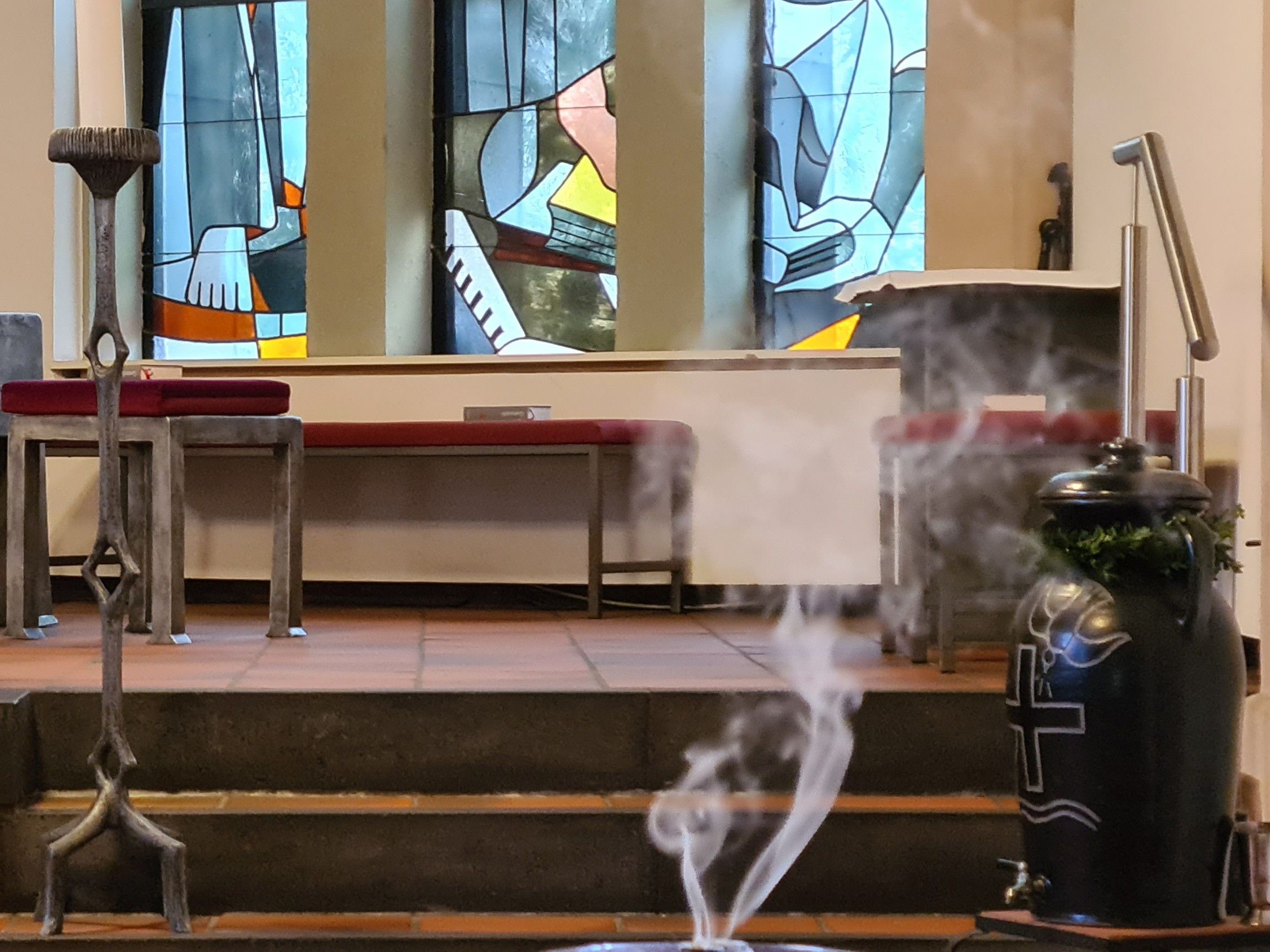 Weihrauchschale auf einem kleinen Tisch vor den Stufen zum Altarraum. Im Hintergrund bunte schmale Fenster. Links im Alttarraum eine Kerze. Von der Weihrauchschale steigt Racu in zwei schwingenden Fäden auf.