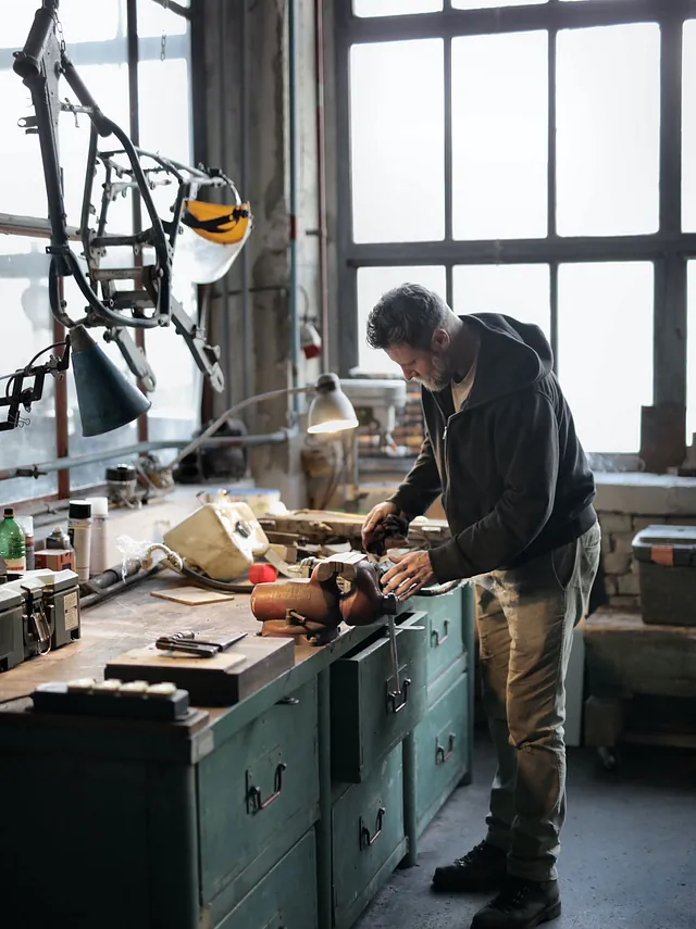 In einer klassichen Werkstatt für Holzarbeit steht ein Mann konzentriert an einer Werkbank und arbeitet behutsam an einem Werkstück. Große Fenster im Hintergrund. Viel Material auf Ablagen.